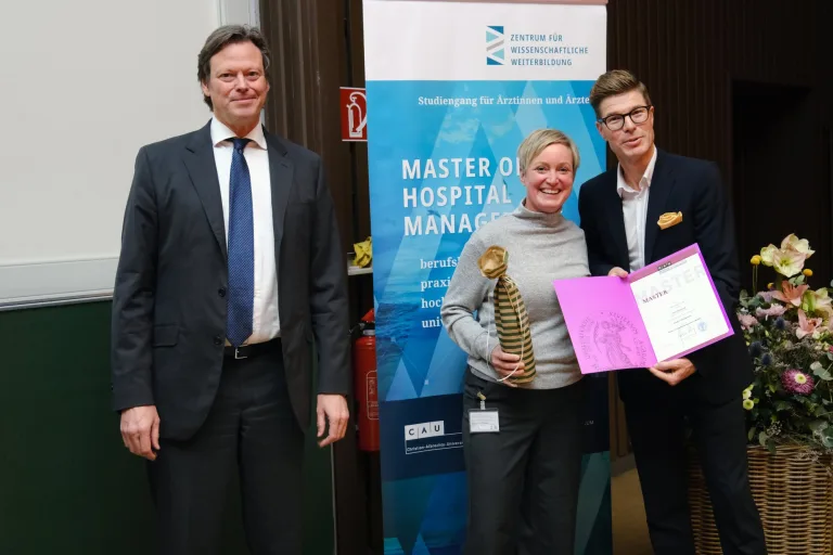 Absolventin mit Urkunde und Geschenk zwischen zwei Männern vor dem Banner Master of Hospital Management