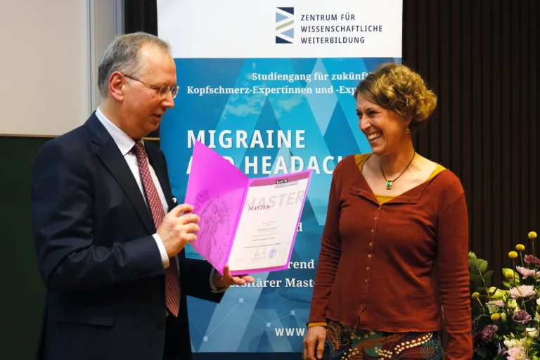 Übergabe einer Masterurkunde vor dem Banner des Studiengangs Migraine and Headache Medicine