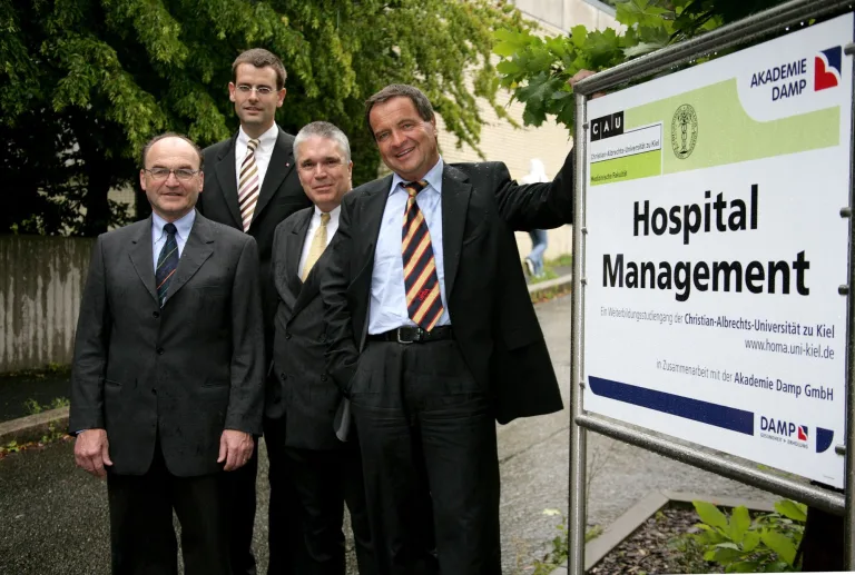 Vier Männer im Anzug stehen neben dem ersten Schild „Hospital Management“